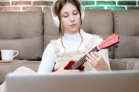 Young caucasian hipster woman is engaged in online ukulele lesson at home, sitting on the floor, on...