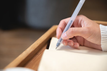 Close-up of female hands writing with gel black pen in leather notepad on wooden table at homeの写真素材