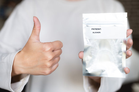 female hands holding a silver package with plastic braces inside, showing a thumbs up gesture, against a blurred backgroundの写真素材