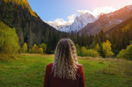 rear view, a blonde woman in a red coat stands against the backdrop of snow-capped mountains and a dense green forestの写真素材