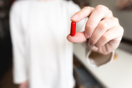 hand holds a capsule or tablet close-up. The hand of a woman holding a medicine in a red capsule with her fingers against the background of herself. Healthy lifestyleの写真素材