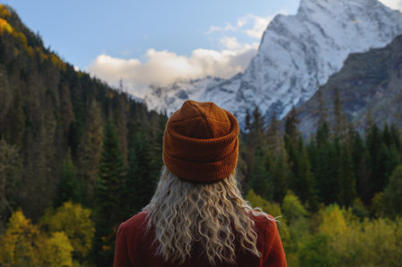 Blonde woman standing with her back to the camera and watching magical clouds in a mountainous area. Woman in a hat in summer or autumn in the snowy mountains next to the green forestの写真素材