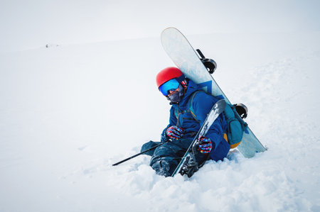 man with ski equipment and a snowboard is sitting a snowy mountain. Blue sky and snowy mountain in the background.の写真素材