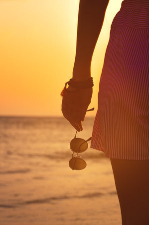 Hand holds stylish round sunglasses at sunset. Summer atmosphere near the coast of the sea, a woman on a walk holds glasses in her hand, close-upの写真素材