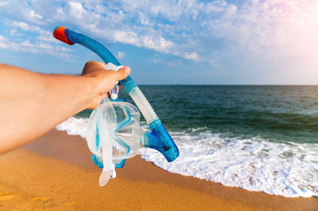 man holds a snorkeling mask in his hand against a background of a blue sky with clouds and a turquoise ocean. Diving mask in a mans hand. Photo of mask and snorkel for swimming in the seaの写真素材
