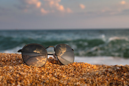 On the beach, sunglasses lie on the sand in the background a wave rolls from the sea. Close-up of leisure accessory, no peopleの写真素材