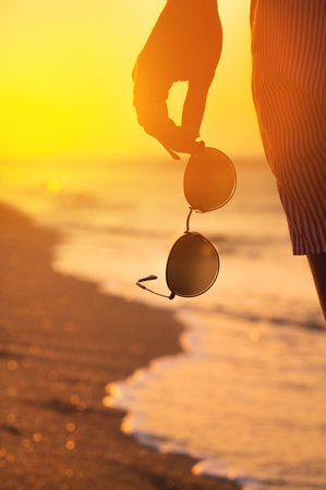 Close-up of a womans hand holding stylish round sunglasses at sunset. Summer atmosphere near the sea coast, a woman on a walk holds glasses in her hand. Vertical shotの写真素材