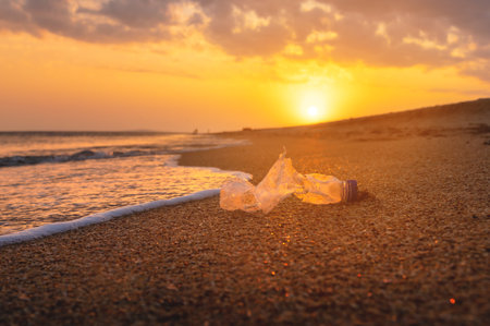 Close-up of environmental and sea pollution concept. Ocean pollution from plastic waste. An old torn bottle lies on wet golden sand at sunsetの写真素材