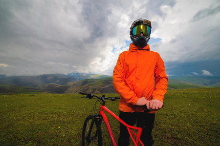 cyclist in an orange bright jacket and a protective helmet stands and holds a bicycle in the mountains, outside the city, off the road in summer in cloudy weatherの写真素材