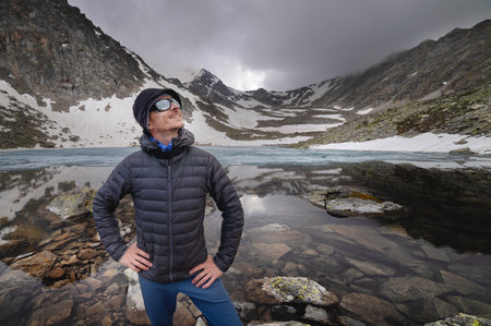 young tourist stands on the top of a mountain against the backdrop of a mountain lake. Happy guy...