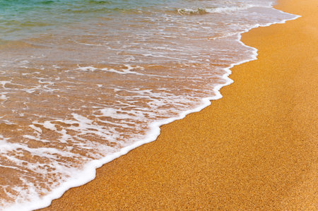 Background image of a soft blue ocean wave on a sandy beach. Close-up sea wave with white foam rolls onto golden sandの写真素材