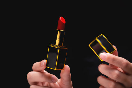 womans hand holding a red lipstick in a gold-black frame in one hand, in the other hand a lipstick cap, close-up on a black backgroundの写真素材