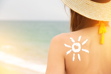 Sun protection. A beautiful woman applied sunscreen to her tanned shoulder in the shape of a sun. Skin care. A girl on the beach sits with her back to the camera in a straw hatの写真素材
