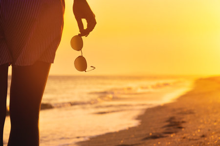 woman in shorts stands on the shore with sunglasses in her hand, close-up. Warm colors, beach holiday at sunsetの写真素材