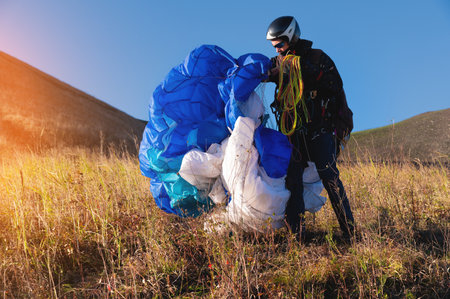 A young male paraglider at sunset in the mountains collects his paraglider to take it to the starting point. Paragliding sportの写真素材