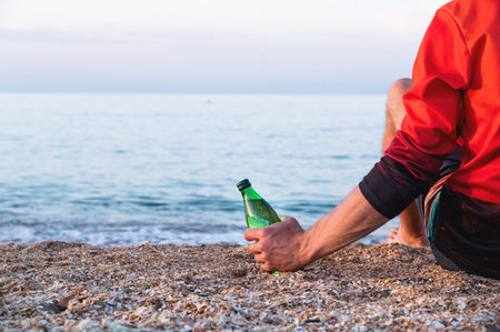 A man in a red jacket sits on the beach in the morning before sunrise with a glass bottle in his hand. Alcoholism on the beachの写真素材