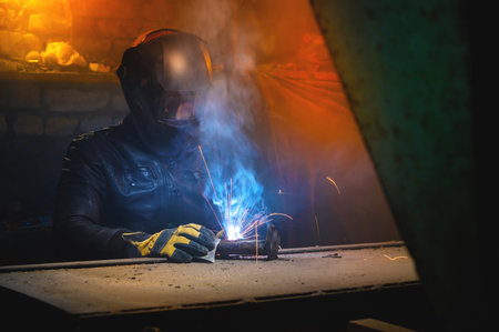 A welder in a leather jacket and welding mask welds in a dark room.