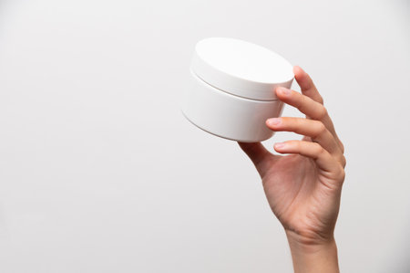 Close-up of a womans hand holding a white jar of cream, mockup for advertising. Girl on a white background showing a cosmetic productの写真素材