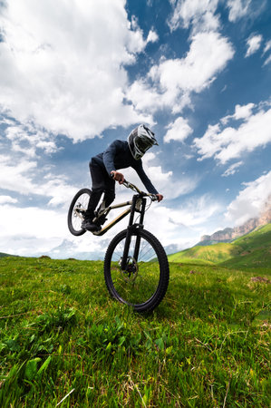 mountain biker takes off with his rear wheel leaning on his front wheel, performing a manuel or wheelie trick, against a mountain landscape in the fields in summerの写真素材