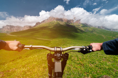 Mountain bike descending from a hill, view from the eyes of a cyclist. First person view of male hands on a mtb bike against the backdrop of rocky mountains wrapped in fluffy cloudsの写真素材