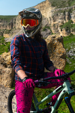 Portrait of a young man in a protective helmet and gloves, in bright clothes, sitting on a downhill bike and looking into the distance. Extreme rest in the mountainsの写真素材
