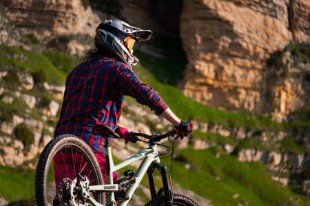 Unrecognizable athlete in protective gear standing in a field and looking at the camera against the backdrop mountain rock. MTB sports concept.の写真素材