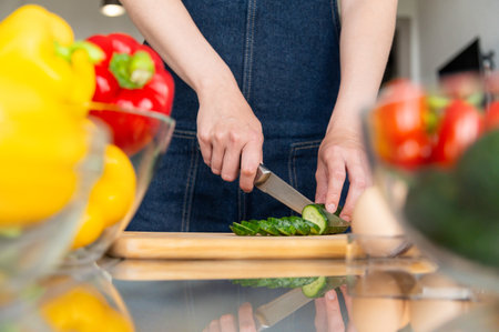 Unrecognizable housewife and her hands cutting green fresh cucumber on wooden cutting board among vegetables and prepared ingredientsの写真素材
