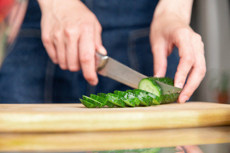 Close-up of knife cutting juicy bright cucumber on wooden cutting board. Preparing ingredients for cooking. Healthy and wholesome food conceptの写真素材