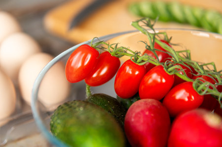 Cherry tomatoes and radishes in a glass bowl in the foreground on a kitchen table, in the background, out of focus, a dozen chicken eggs and sliced cucumber on a cutting boardの写真素材