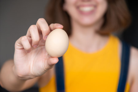 womans hand holds a raw chicken egg against the background of herself and her smile. Portrait of an unrecognizable girl showing the product from which she will prepare breakfastの写真素材