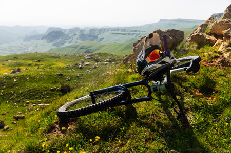 Mountain bike with helmet and mask lying on grass high in mountains against mountain landscape of...