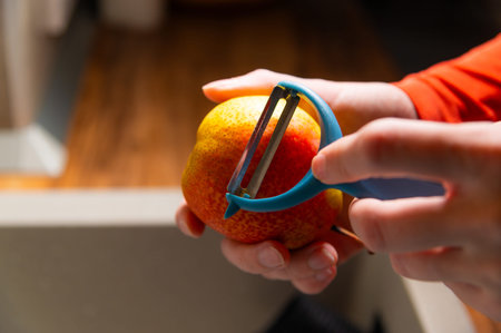 Person peeling a red pear with a blue vegetable peeler in a kitchen setting, wooden floor and blurred background elements visibleの写真素材
