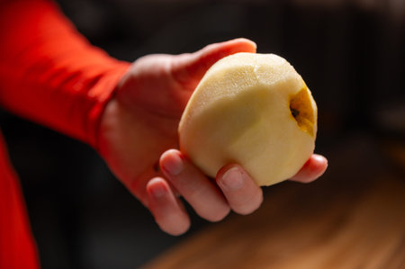 Hand holding a peeled pear with a partially visible brown spot, wearing a red long-sleeve shirt, wooden surface in the background with soft lightingの写真素材