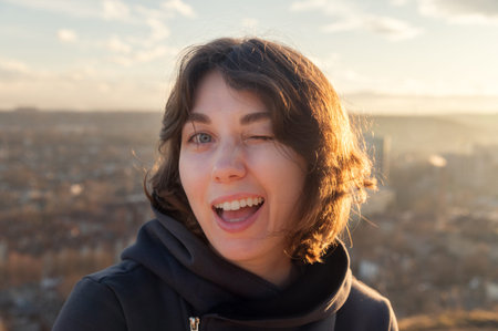 Young woman with short brown hair winks playfully at the camera, wearing a black hoodie, with a scenic city landscape and cloudy sky in the backgroundの写真素材