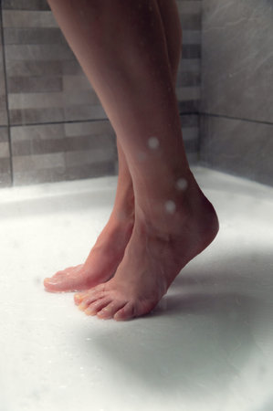 Female Close-up of bare feet standing on wet shower floor with water droplets and tiled wall in the...