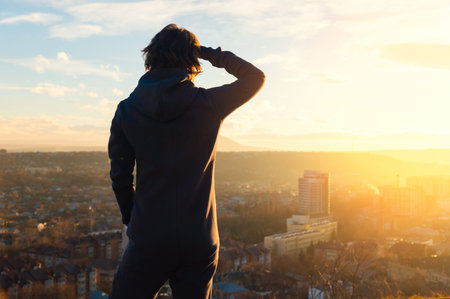 Female figure in a fleece jumpsuit stands on hilltop overlooking cityscape at sunset, with buildings...