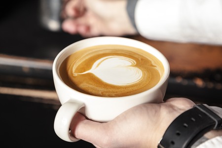 A professional Barista holds a Cup of coffee, making a beautiful heart shape latte art. Hot art latte coffee with heart shape in white Cupの写真素材