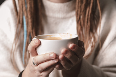 Close-up of a white Cup of hot latte art coffee with a heart shape in the hands of a young girl. Woman wearing warm winter knitted white sweater. Toned. Coffee Girl Concept Relaxation Coffee Mug.の写真素材