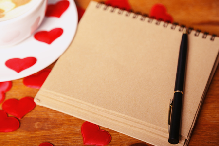 Valentine's day background. A white cappuccino Cup with a heart-shaped latte art, a notebook with vintage sheets of paper and a pen on a wooden background with scattered soft silk hearts. Copy spaceの写真素材