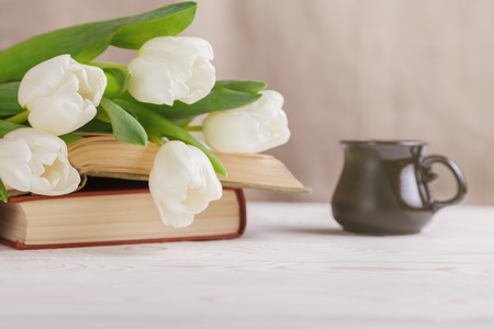 Beautiful composition with white tulips and old books in the foreground, and a Cup of coffee in the distance, on a beige blurred background in the morning light. Spring, reading, coffee breakの写真素材