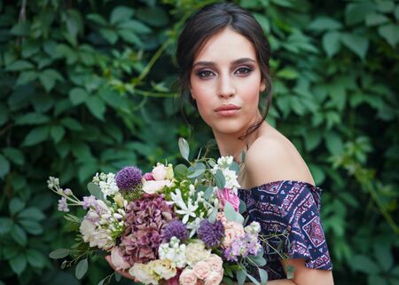 Portrait of a beautiful young woman with perfect makeup and hairstyle while holding a bouquet of wildflowers. Standing against the ivy wall. The concept of happiness and loveの写真素材