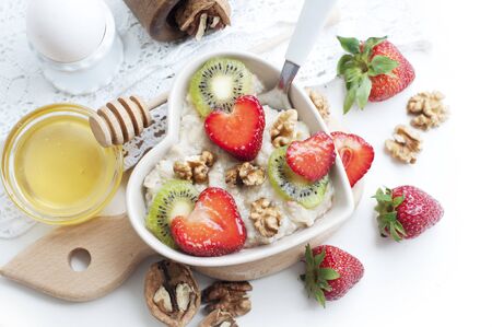 Breakfast with oatmeal with strawberries, kiwi, honey, walnuts and egg on white background. Breakfast for child. Oatmeal in a white heart-shaped plate. Top viewの写真素材