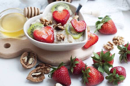 Breakfast with oatmeal with strawberries, kiwi, honey, walnuts and egg on white background. Breakfast for child. Oatmeal in a white heart-shaped plateの写真素材