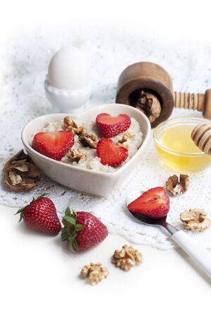 Breakfast with oatmeal with strawberries, honey, walnuts and egg on white background. Breakfast for child. Oatmeal in a white heart-shaped bowlの写真素材