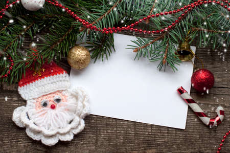 New Year and Christmas card with handmade Santa Claus, shine balls, bell and fir branches on a wooden background. A postcard with Christmas decorations and white paper.の写真素材