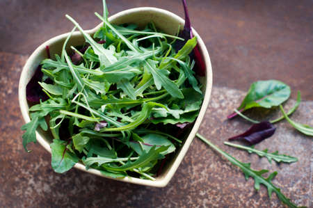 Fresh arugula and spinach in a heart shaped bowl on dark brown grange background. Top viewの写真素材