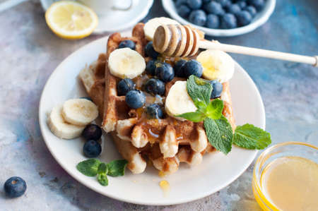 Belgian waffles with blueberries, bananas, honey and mint on a white plate. Breakfast with a cup of tea with a lemon and sweet waffles.の写真素材