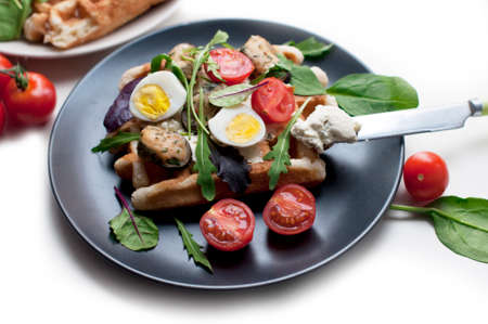 Belgian waffles with mussels, tomatoes, egg and green salad, arugula, spinach on black plate on white background. Cream cheese on the knifeの写真素材
