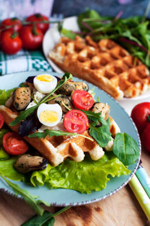 Belgian waffles with mussels, tomatoes, egg and green salad, arugula, spinach on blue plate on white backgroundの写真素材