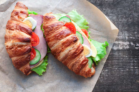 Croissant sandwich with ham, cheese, fresh salad, tomatoes, eggs and cucumber on wooden background. Breakfast with fresh croissant sandwich. Top viewの写真素材
